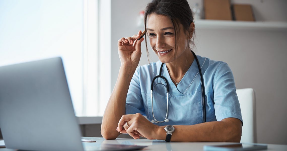 Charming young woman physician sitting at the table with laptop and smiling while giving online consultation