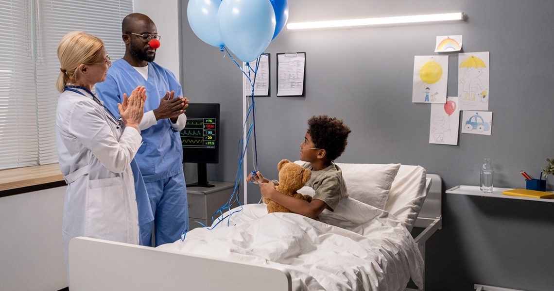 Two doctors clapping hands and having fun the little boy while he sitting in bed with toy and balloons, they congratulating him with recovering